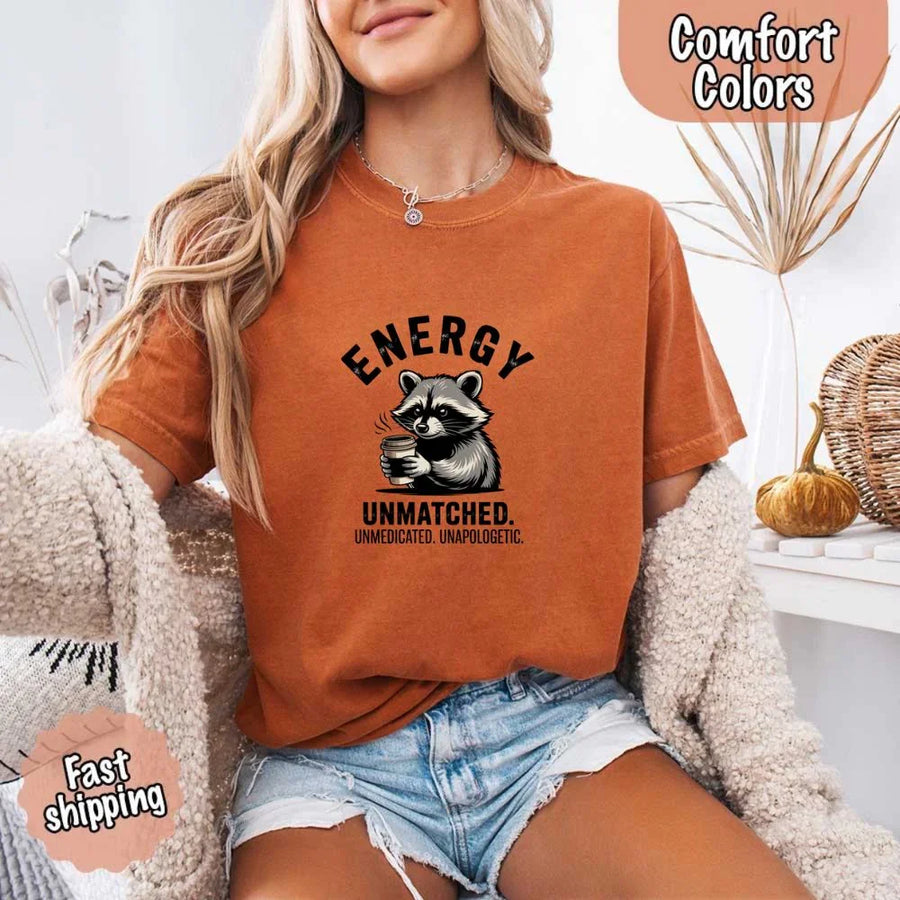 May include a black and white raccoon illustration with a tired, unbothered expression, holding a coffee cup, and bold text that reads “ENERGY UNMATCHED. UNMEDICATED. UNAPOLOGETIC.” conveying raw, unapologetic humor and mood.
