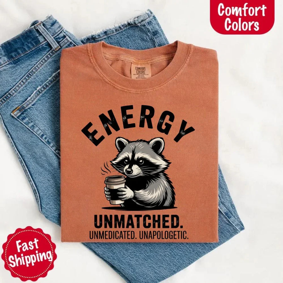 May include a black and white raccoon illustration with a tired, unbothered expression, holding a coffee cup, and bold text that reads “ENERGY UNMATCHED. UNMEDICATED. UNAPOLOGETIC.” conveying raw, unapologetic humor and mood.