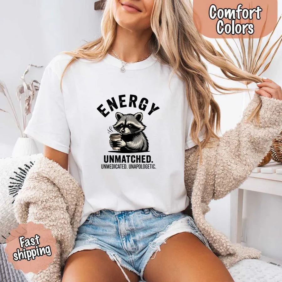May include a black and white raccoon illustration with a tired, unbothered expression, holding a coffee cup, and bold text that reads “ENERGY UNMATCHED. UNMEDICATED. UNAPOLOGETIC.” conveying raw, unapologetic humor and mood.