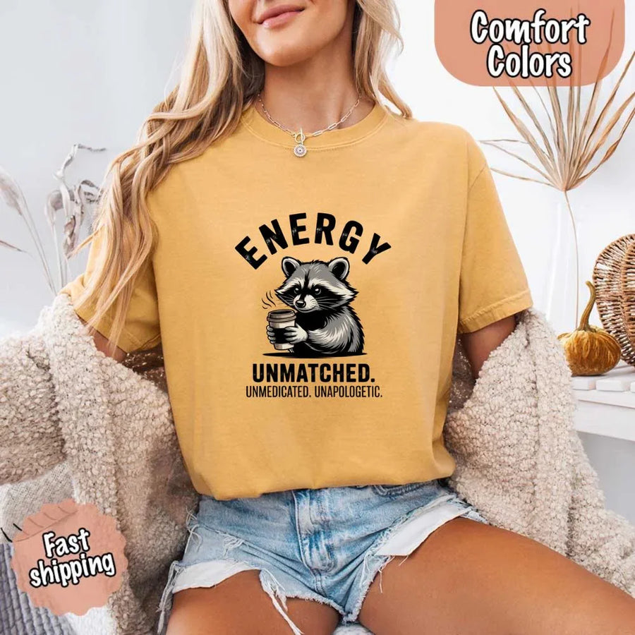 May include a black and white raccoon illustration with a tired, unbothered expression, holding a coffee cup, and bold text that reads “ENERGY UNMATCHED. UNMEDICATED. UNAPOLOGETIC.” conveying raw, unapologetic humor and mood.