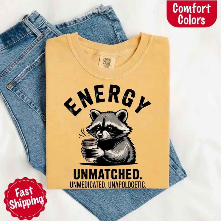 May include a black and white raccoon illustration with a tired, unbothered expression, holding a coffee cup, and bold text that reads “ENERGY UNMATCHED. UNMEDICATED. UNAPOLOGETIC.” conveying raw, unapologetic humor and mood.