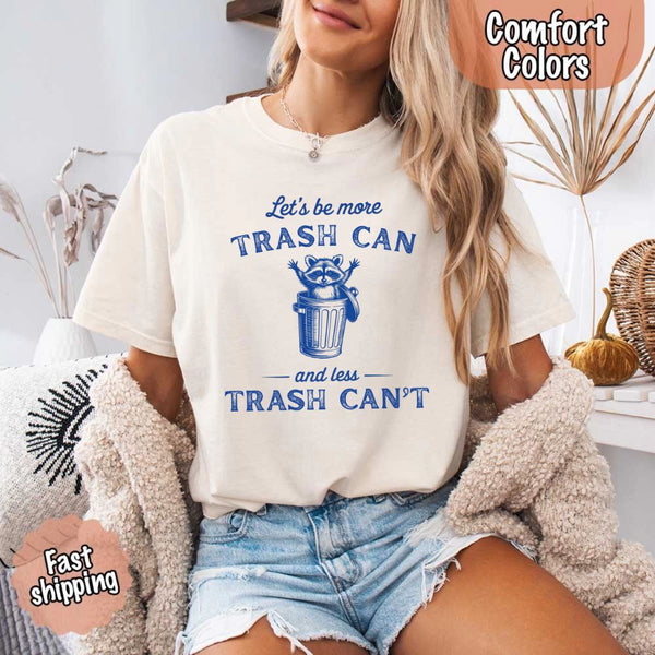 May Include: Comfort Colors t-shirt with a vintage-style blue graphic that features a cheerful raccoon popping out of a trash can with its arms raised. The design includes playful, curved typography that reads 'Let’s be more TRASH CAN' above the raccoon and 'and less TRASH CAN’T' below it. The raccoon has a big smile and cartoon eyes, giving the shirt a humorous and uplifting feel. Ideal for motivational, funny, or animal-themed apparel collections