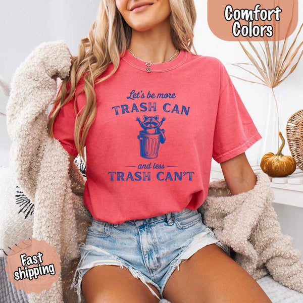 May Include: Comfort Colors t-shirt with a vintage-style blue graphic that features a cheerful raccoon popping out of a trash can with its arms raised. The design includes playful, curved typography that reads 'Let’s be more TRASH CAN' above the raccoon and 'and less TRASH CAN’T' below it. The raccoon has a big smile and cartoon eyes, giving the shirt a humorous and uplifting feel. Ideal for motivational, funny, or animal-themed apparel collections