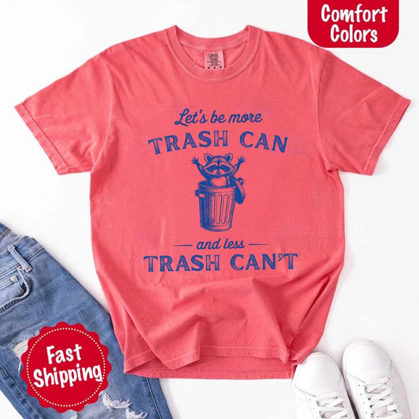 May Include: Comfort Colors t-shirt with a vintage-style blue graphic that features a cheerful raccoon popping out of a trash can with its arms raised. The design includes playful, curved typography that reads 'Let’s be more TRASH CAN' above the raccoon and 'and less TRASH CAN’T' below it. The raccoon has a big smile and cartoon eyes, giving the shirt a humorous and uplifting feel. Ideal for motivational, funny, or animal-themed apparel collections