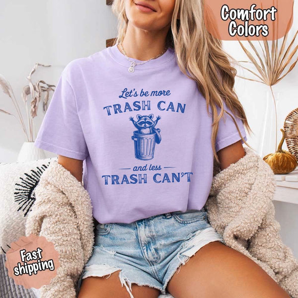 May Include: Comfort Colors t-shirt with a vintage-style blue graphic that features a cheerful raccoon popping out of a trash can with its arms raised. The design includes playful, curved typography that reads 'Let’s be more TRASH CAN' above the raccoon and 'and less TRASH CAN’T' below it. The raccoon has a big smile and cartoon eyes, giving the shirt a humorous and uplifting feel. Ideal for motivational, funny, or animal-themed apparel collections