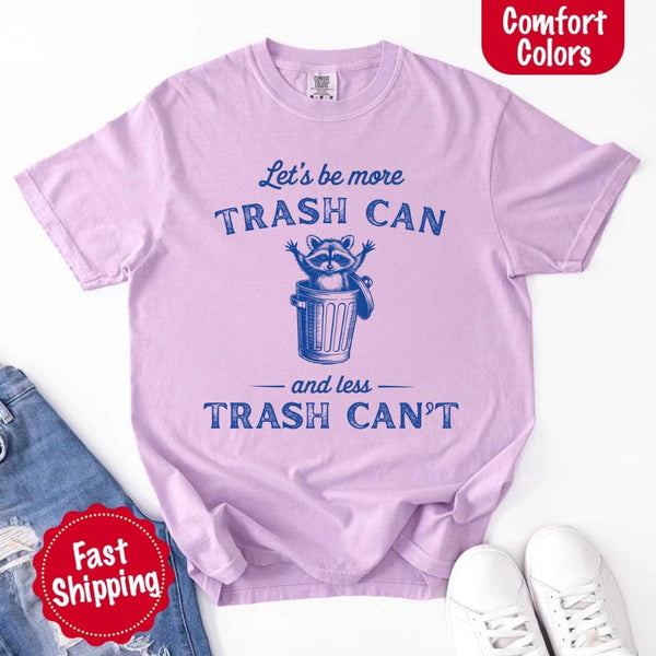 May Include: Comfort Colors t-shirt with a vintage-style blue graphic that features a cheerful raccoon popping out of a trash can with its arms raised. The design includes playful, curved typography that reads 'Let’s be more TRASH CAN' above the raccoon and 'and less TRASH CAN’T' below it. The raccoon has a big smile and cartoon eyes, giving the shirt a humorous and uplifting feel. Ideal for motivational, funny, or animal-themed apparel collections