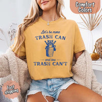 May Include: Comfort Colors t-shirt with a vintage-style blue graphic that features a cheerful raccoon popping out of a trash can with its arms raised. The design includes playful, curved typography that reads 'Let’s be more TRASH CAN' above the raccoon and 'and less TRASH CAN’T' below it. The raccoon has a big smile and cartoon eyes, giving the shirt a humorous and uplifting feel. Ideal for motivational, funny, or animal-themed apparel collections
