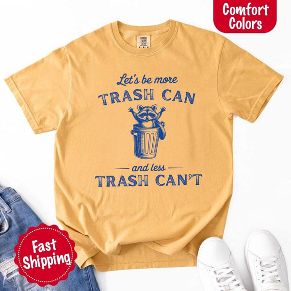 May Include: Comfort Colors t-shirt with a vintage-style blue graphic that features a cheerful raccoon popping out of a trash can with its arms raised. The design includes playful, curved typography that reads 'Let’s be more TRASH CAN' above the raccoon and 'and less TRASH CAN’T' below it. The raccoon has a big smile and cartoon eyes, giving the shirt a humorous and uplifting feel. Ideal for motivational, funny, or animal-themed apparel collections