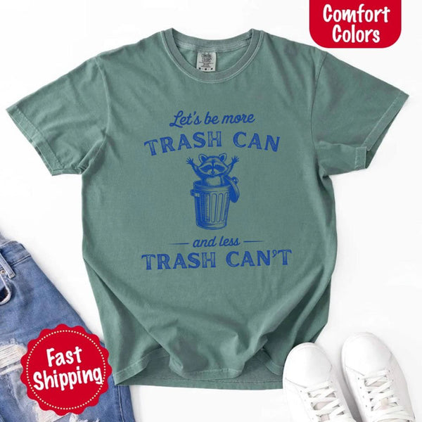 May Include: Comfort Colors t-shirt with a vintage-style blue graphic that features a cheerful raccoon popping out of a trash can with its arms raised. The design includes playful, curved typography that reads 'Let’s be more TRASH CAN' above the raccoon and 'and less TRASH CAN’T' below it. The raccoon has a big smile and cartoon eyes, giving the shirt a humorous and uplifting feel. Ideal for motivational, funny, or animal-themed apparel collections