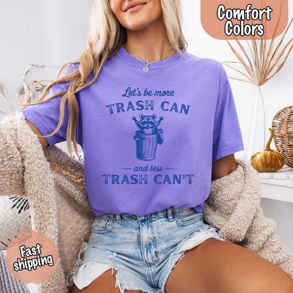 May Include: Comfort Colors t-shirt with a vintage-style blue graphic that features a cheerful raccoon popping out of a trash can with its arms raised. The design includes playful, curved typography that reads 'Let’s be more TRASH CAN' above the raccoon and 'and less TRASH CAN’T' below it. The raccoon has a big smile and cartoon eyes, giving the shirt a humorous and uplifting feel. Ideal for motivational, funny, or animal-themed apparel collections