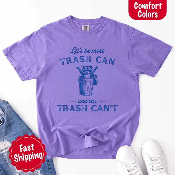 May Include: Comfort Colors t-shirt with a vintage-style blue graphic that features a cheerful raccoon popping out of a trash can with its arms raised. The design includes playful, curved typography that reads 'Let’s be more TRASH CAN' above the raccoon and 'and less TRASH CAN’T' below it. The raccoon has a big smile and cartoon eyes, giving the shirt a humorous and uplifting feel. Ideal for motivational, funny, or animal-themed apparel collections