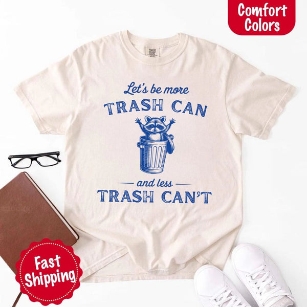 May Include: Comfort Colors t-shirt with a vintage-style blue graphic that features a cheerful raccoon popping out of a trash can with its arms raised. The design includes playful, curved typography that reads 'Let’s be more TRASH CAN' above the raccoon and 'and less TRASH CAN’T' below it. The raccoon has a big smile and cartoon eyes, giving the shirt a humorous and uplifting feel. Ideal for motivational, funny, or animal-themed apparel collections