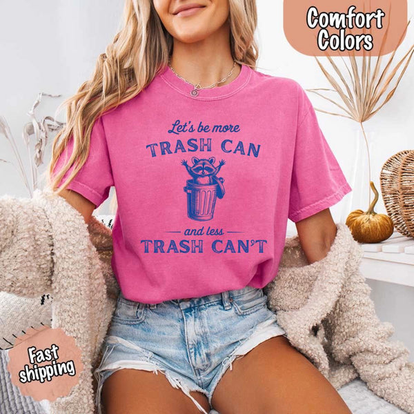 May Include: Comfort Colors t-shirt with a vintage-style blue graphic that features a cheerful raccoon popping out of a trash can with its arms raised. The design includes playful, curved typography that reads 'Let’s be more TRASH CAN' above the raccoon and 'and less TRASH CAN’T' below it. The raccoon has a big smile and cartoon eyes, giving the shirt a humorous and uplifting feel. Ideal for motivational, funny, or animal-themed apparel collections