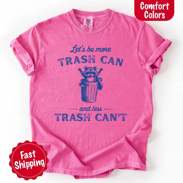 May Include: Comfort Colors t-shirt with a vintage-style blue graphic that features a cheerful raccoon popping out of a trash can with its arms raised. The design includes playful, curved typography that reads 'Let’s be more TRASH CAN' above the raccoon and 'and less TRASH CAN’T' below it. The raccoon has a big smile and cartoon eyes, giving the shirt a humorous and uplifting feel. Ideal for motivational, funny, or animal-themed apparel collections