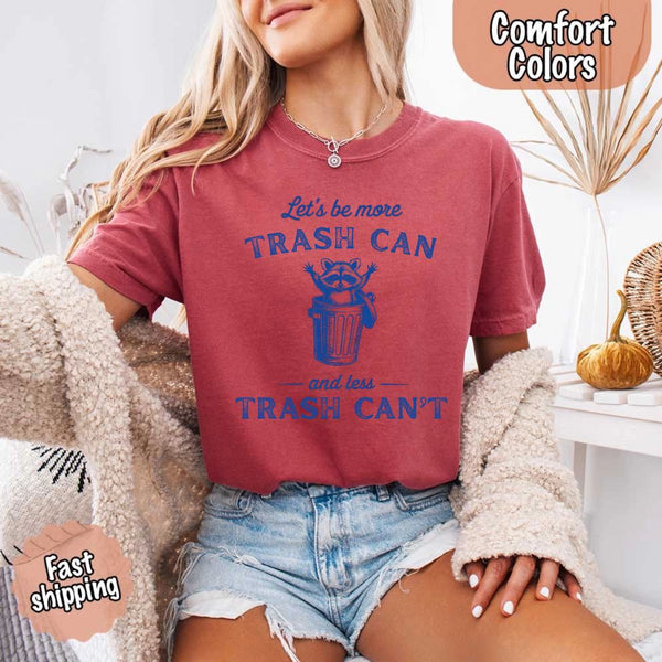 May Include: Comfort Colors t-shirt with a vintage-style blue graphic that features a cheerful raccoon popping out of a trash can with its arms raised. The design includes playful, curved typography that reads 'Let’s be more TRASH CAN' above the raccoon and 'and less TRASH CAN’T' below it. The raccoon has a big smile and cartoon eyes, giving the shirt a humorous and uplifting feel. Ideal for motivational, funny, or animal-themed apparel collections