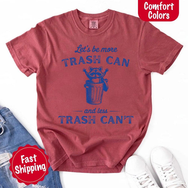 May Include: Comfort Colors t-shirt with a vintage-style blue graphic that features a cheerful raccoon popping out of a trash can with its arms raised. The design includes playful, curved typography that reads 'Let’s be more TRASH CAN' above the raccoon and 'and less TRASH CAN’T' below it. The raccoon has a big smile and cartoon eyes, giving the shirt a humorous and uplifting feel. Ideal for motivational, funny, or animal-themed apparel collections