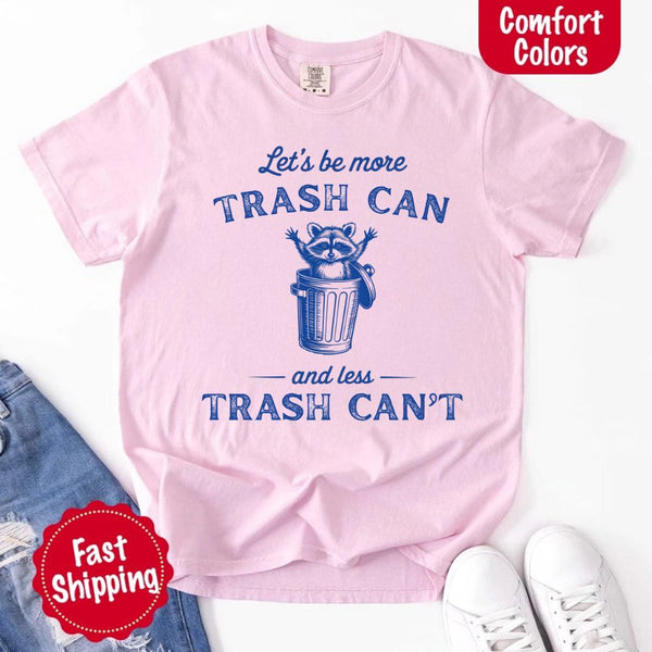 May Include: Comfort Colors t-shirt with a vintage-style blue graphic that features a cheerful raccoon popping out of a trash can with its arms raised. The design includes playful, curved typography that reads 'Let’s be more TRASH CAN' above the raccoon and 'and less TRASH CAN’T' below it. The raccoon has a big smile and cartoon eyes, giving the shirt a humorous and uplifting feel. Ideal for motivational, funny, or animal-themed apparel collections