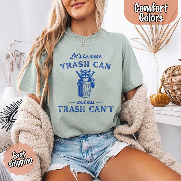 May Include: Comfort Colors t-shirt with a vintage-style blue graphic that features a cheerful raccoon popping out of a trash can with its arms raised. The design includes playful, curved typography that reads 'Let’s be more TRASH CAN' above the raccoon and 'and less TRASH CAN’T' below it. The raccoon has a big smile and cartoon eyes, giving the shirt a humorous and uplifting feel. Ideal for motivational, funny, or animal-themed apparel collections