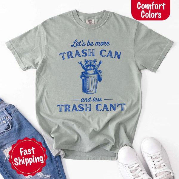 May Include: Comfort Colors t-shirt with a vintage-style blue graphic that features a cheerful raccoon popping out of a trash can with its arms raised. The design includes playful, curved typography that reads 'Let’s be more TRASH CAN' above the raccoon and 'and less TRASH CAN’T' below it. The raccoon has a big smile and cartoon eyes, giving the shirt a humorous and uplifting feel. Ideal for motivational, funny, or animal-themed apparel collections
