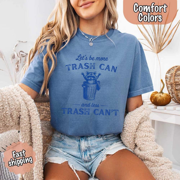 May Include: Comfort Colors t-shirt with a vintage-style blue graphic that features a cheerful raccoon popping out of a trash can with its arms raised. The design includes playful, curved typography that reads 'Let’s be more TRASH CAN' above the raccoon and 'and less TRASH CAN’T' below it. The raccoon has a big smile and cartoon eyes, giving the shirt a humorous and uplifting feel. Ideal for motivational, funny, or animal-themed apparel collections
