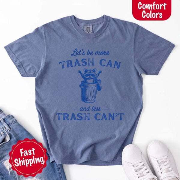 May Include: Comfort Colors t-shirt with a vintage-style blue graphic that features a cheerful raccoon popping out of a trash can with its arms raised. The design includes playful, curved typography that reads 'Let’s be more TRASH CAN' above the raccoon and 'and less TRASH CAN’T' below it. The raccoon has a big smile and cartoon eyes, giving the shirt a humorous and uplifting feel. Ideal for motivational, funny, or animal-themed apparel collections