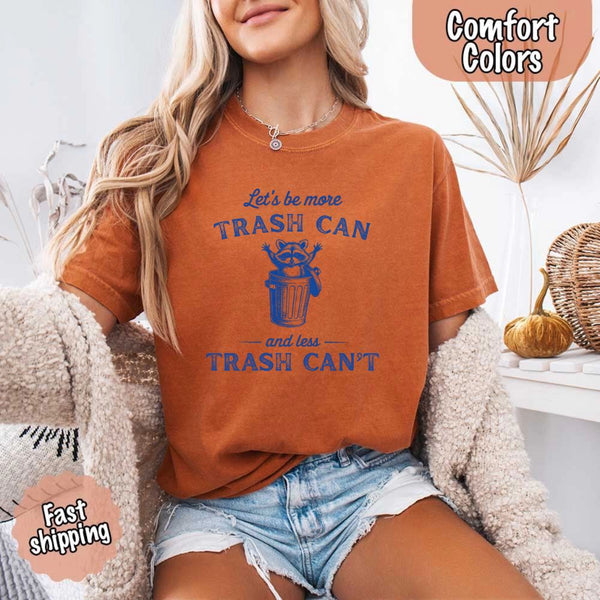 May Include: Comfort Colors t-shirt with a vintage-style blue graphic that features a cheerful raccoon popping out of a trash can with its arms raised. The design includes playful, curved typography that reads 'Let’s be more TRASH CAN' above the raccoon and 'and less TRASH CAN’T' below it. The raccoon has a big smile and cartoon eyes, giving the shirt a humorous and uplifting feel. Ideal for motivational, funny, or animal-themed apparel collections