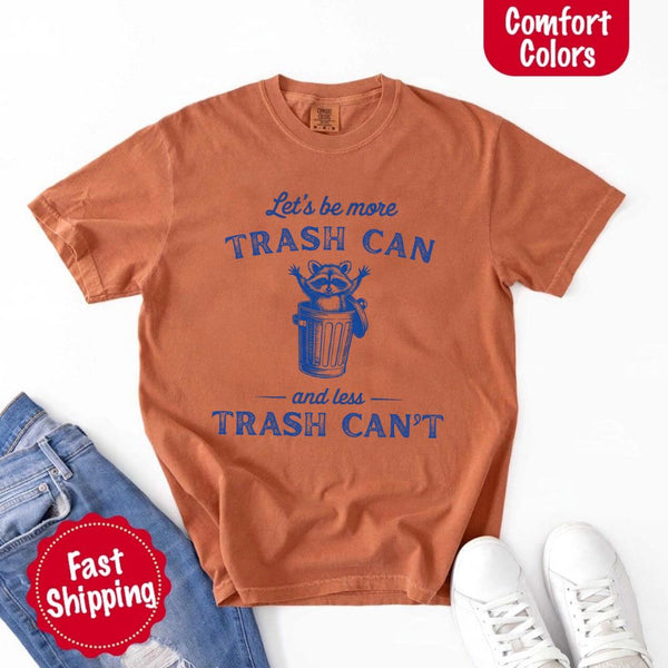 May Include: Comfort Colors t-shirt with a vintage-style blue graphic that features a cheerful raccoon popping out of a trash can with its arms raised. The design includes playful, curved typography that reads 'Let’s be more TRASH CAN' above the raccoon and 'and less TRASH CAN’T' below it. The raccoon has a big smile and cartoon eyes, giving the shirt a humorous and uplifting feel. Ideal for motivational, funny, or animal-themed apparel collections