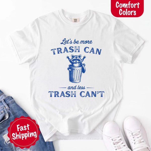 May Include: Comfort Colors t-shirt with a vintage-style blue graphic that features a cheerful raccoon popping out of a trash can with its arms raised. The design includes playful, curved typography that reads 'Let’s be more TRASH CAN' above the raccoon and 'and less TRASH CAN’T' below it. The raccoon has a big smile and cartoon eyes, giving the shirt a humorous and uplifting feel. Ideal for motivational, funny, or animal-themed apparel collections