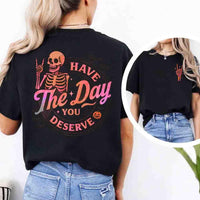 May include skeleton Halloween graphic with holographic lettering ‘Have the Day You Deserve,’ skeleton hand gesture, bats, spider webs, and pumpkin accent – spooky graphic tee design.