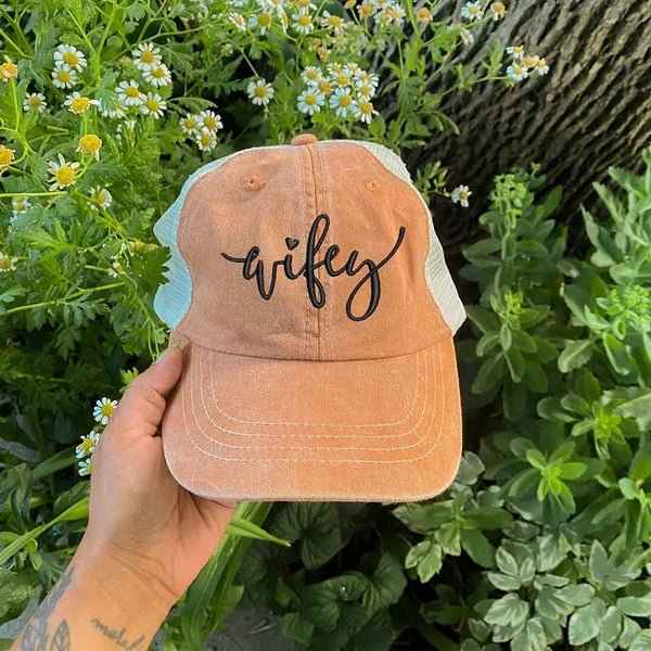 May include an embroidered pigment-dyed trucker hat with mesh back, featuring “Wifey” or “Hubby” text, designed as a casual and stylish gift for spouses or anniversaries.
