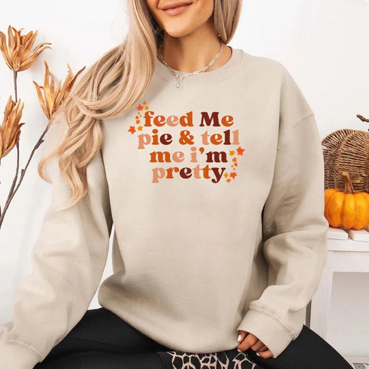 May include a cozy sweatshirt displayed with jeans and fall-themed decor, featuring retro-style text that reads “feed me pie & tell me i’m pretty” in warm autumn shades of orange and brown, accented with small leaf graphics for a seasonal touch.