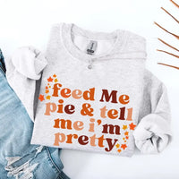 May include a cozy sweatshirt displayed with jeans and fall-themed decor, featuring retro-style text that reads “feed me pie & tell me i’m pretty” in warm autumn shades of orange and brown, accented with small leaf graphics for a seasonal touch.