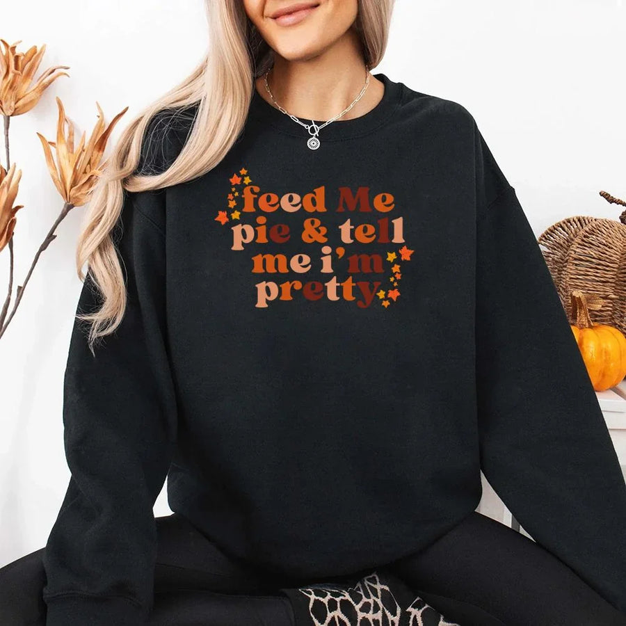 May include a cozy sweatshirt displayed with jeans and fall-themed decor, featuring retro-style text that reads “feed me pie & tell me i’m pretty” in warm autumn shades of orange and brown, accented with small leaf graphics for a seasonal touch.