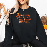 May include a cozy sweatshirt displayed with jeans and fall-themed decor, featuring retro-style text that reads “feed me pie & tell me i’m pretty” in warm autumn shades of orange and brown, accented with small leaf graphics for a seasonal touch.