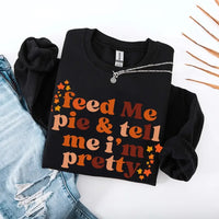 May include a cozy sweatshirt displayed with jeans and fall-themed decor, featuring retro-style text that reads “feed me pie & tell me i’m pretty” in warm autumn shades of orange and brown, accented with small leaf graphics for a seasonal touch.