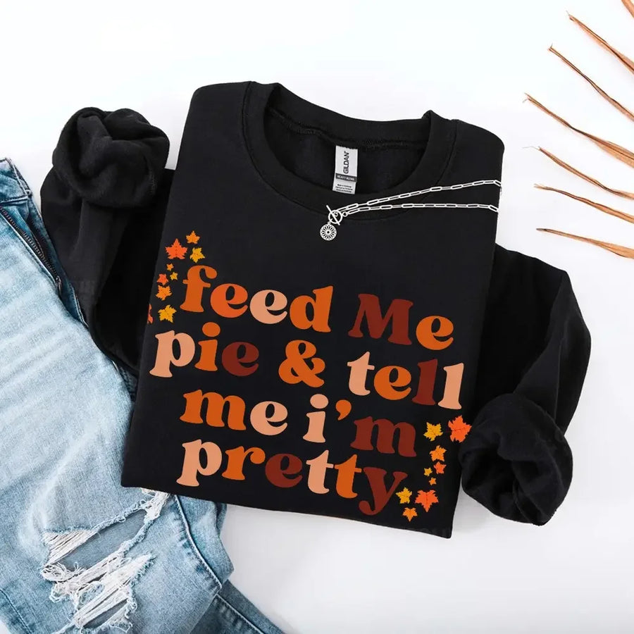 May include a cozy sweatshirt displayed with jeans and fall-themed decor, featuring retro-style text that reads “feed me pie & tell me i’m pretty” in warm autumn shades of orange and brown, accented with small leaf graphics for a seasonal touch.