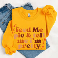 May include a cozy sweatshirt displayed with jeans and fall-themed decor, featuring retro-style text that reads “feed me pie & tell me i’m pretty” in warm autumn shades of orange and brown, accented with small leaf graphics for a seasonal touch.