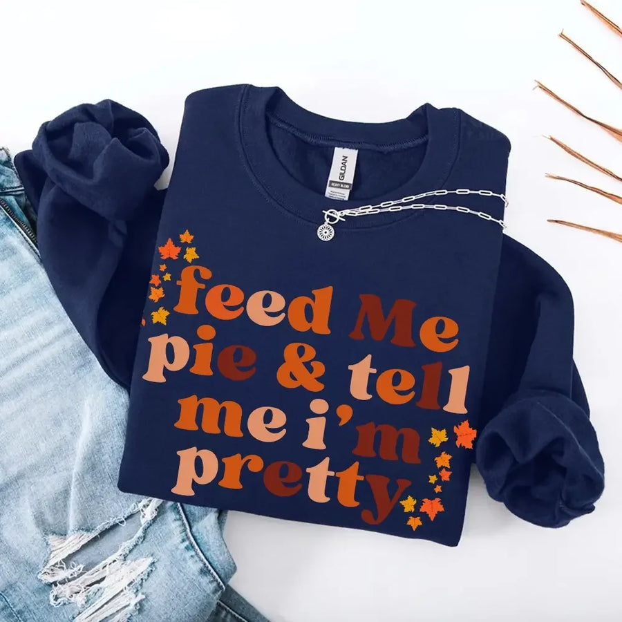 May include a cozy sweatshirt displayed with jeans and fall-themed decor, featuring retro-style text that reads “feed me pie & tell me i’m pretty” in warm autumn shades of orange and brown, accented with small leaf graphics for a seasonal touch.