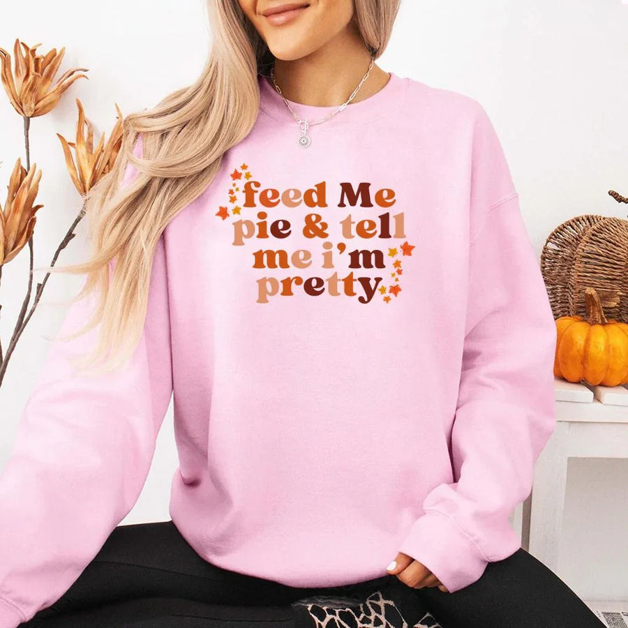 May include a cozy sweatshirt displayed with jeans and fall-themed decor, featuring retro-style text that reads “feed me pie & tell me i’m pretty” in warm autumn shades of orange and brown, accented with small leaf graphics for a seasonal touch.