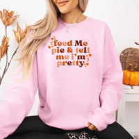 May include a cozy sweatshirt displayed with jeans and fall-themed decor, featuring retro-style text that reads “feed me pie & tell me i’m pretty” in warm autumn shades of orange and brown, accented with small leaf graphics for a seasonal touch.