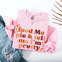 May include a cozy sweatshirt displayed with jeans and fall-themed decor, featuring retro-style text that reads “feed me pie & tell me i’m pretty” in warm autumn shades of orange and brown, accented with small leaf graphics for a seasonal touch.