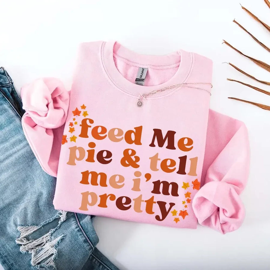 May include a cozy sweatshirt displayed with jeans and fall-themed decor, featuring retro-style text that reads “feed me pie & tell me i’m pretty” in warm autumn shades of orange and brown, accented with small leaf graphics for a seasonal touch.