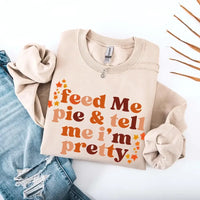 May include a cozy sweatshirt displayed with jeans and fall-themed decor, featuring retro-style text that reads “feed me pie & tell me i’m pretty” in warm autumn shades of orange and brown, accented with small leaf graphics for a seasonal touch.