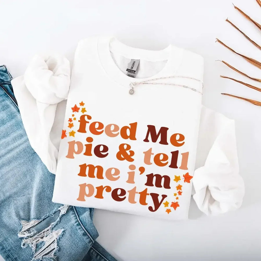 May include a cozy sweatshirt displayed with jeans and fall-themed decor, featuring retro-style text that reads “feed me pie & tell me i’m pretty” in warm autumn shades of orange and brown, accented with small leaf graphics for a seasonal touch.