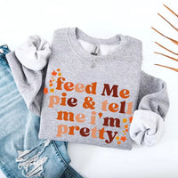 May include a cozy sweatshirt displayed with jeans and fall-themed decor, featuring retro-style text that reads “feed me pie & tell me i’m pretty” in warm autumn shades of orange and brown, accented with small leaf graphics for a seasonal touch.