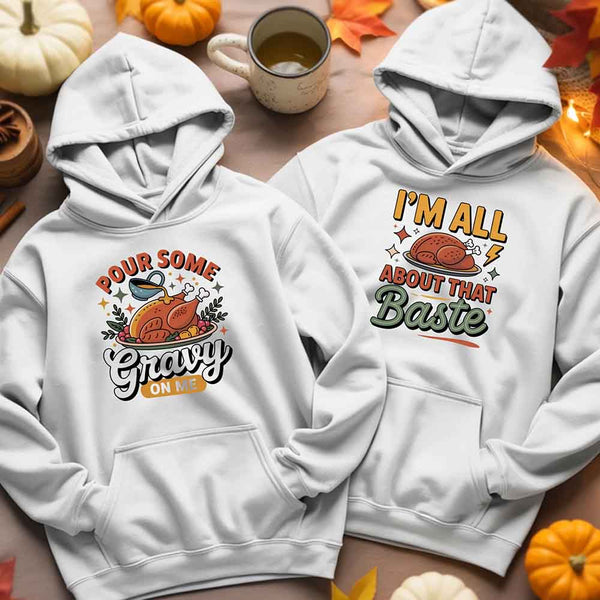 Two white hoodies with Thanksgiving-themed designs on a table with pumpkins and a mug.