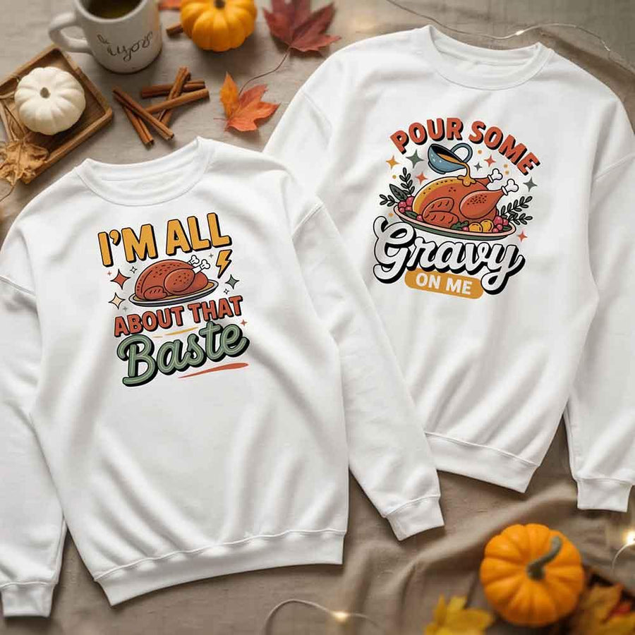 Two white sweatshirts with Thanksgiving-themed graphics on a table with pumpkins and leaves.