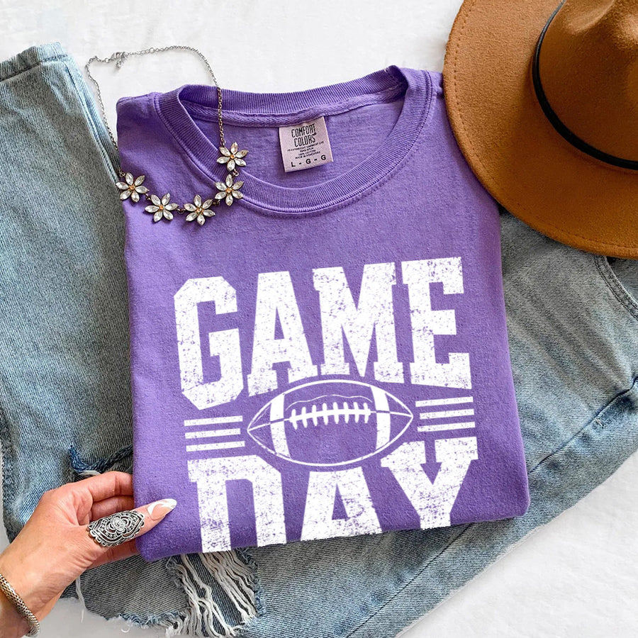 May include a Comfort Colors T-shirt featuring a distressed Game Day graphic with a football design, perfect for football fans, tailgates, watch parties, and sporting events.