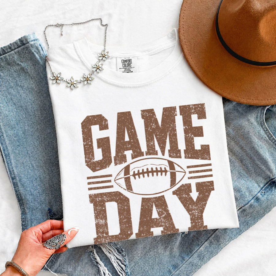 May include a Comfort Colors T-shirt featuring a distressed Game Day graphic with a football design, perfect for football fans, tailgates, watch parties, and sporting events.