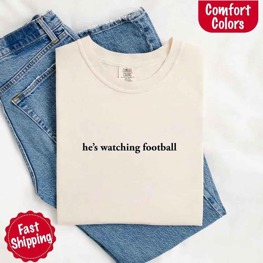 May include a woman wearing an ivory Comfort Colors t-shirt with the phrase 'He’s Watching Football' — styled in a comfy, casual look perfect for Sundays or football season humor
